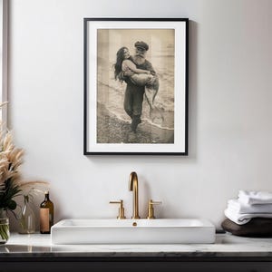 May include: A black and white vintage photograph of a man in a sailor hat holding a mermaid in his arms. The photo is framed in a black frame and hanging on a white wall.