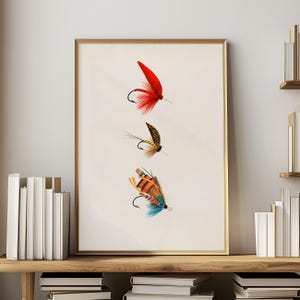 May include: A framed print featuring three detailed illustrations of fishing flies. The flies are in vibrant colors, including red, brown, orange, and blue, with delicate feather and thread details. The artwork is set against a cream-colored background.