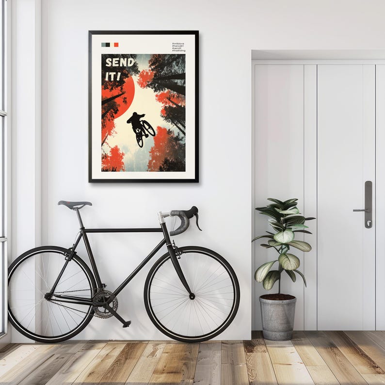May include: A framed poster with the words "SEND IT!" features a mountain biker mid-air against a backdrop of red and black trees and a red sun. A black road bike leans against the wall below the poster. A potted plant sits to the right.