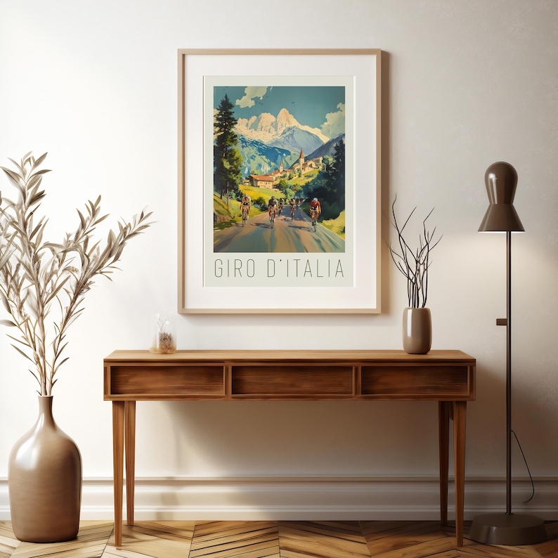 May include: A vintage-style poster featuring a scenic mountain road with cyclists racing up a hill. The poster has a blue sky with white clouds, green trees, and a small village in the background. The text "GIRO D'ITALIA" is printed at the bottom of the poster.