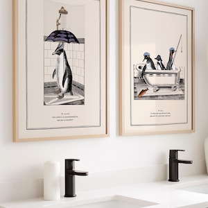 Op de afbeelding: Twee ingelijste kunstprints met pingu&iuml;nillustraties. E&eacute;n toont een pingu&iuml;n onder een paraplu in een douche, de andere toont pingu&iuml;ns in een bad. De prints hebben een beige achtergrond en zitten in licht houten frames.