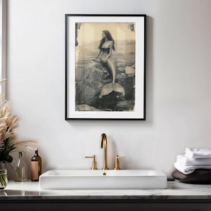 May include: A black and white vintage photograph of a mermaid sitting on a rock in the ocean. The mermaid has long hair and is wearing a seashell bra. The photo is framed in a black frame.