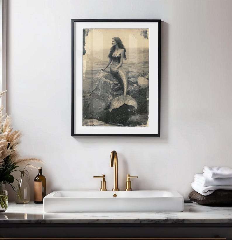 May include: A black and white vintage photograph of a mermaid sitting on a rock in the ocean. The mermaid has long hair and is wearing a seashell bra. The photo is framed in a black frame.