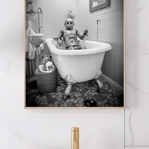 May include: A black and white photograph of a robot taking a bath in a white bathtub with claw feet. The robot is sitting in the tub with its arms crossed. The bathroom is tiled in white and has a mirror on the wall.