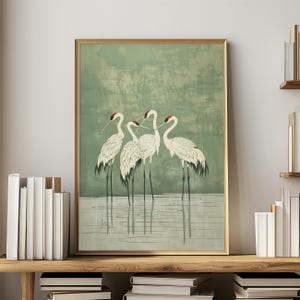Op de afbeelding: Een ingelijste kunstdruk met vier witte kraanvogels met zwarte poten en rode kopmarkeringen, staand in het water. De achtergrond is een gedempt groen met een textuur, en de lijst is goudkleurig. Het kunstwerk wordt tentoongesteld op een houten plank met boeken.