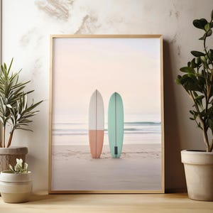 May include: A framed print of two surfboards on a beach. The surfboards are white and light blue with a pink and green stripe. The beach is sandy and the water is blue.