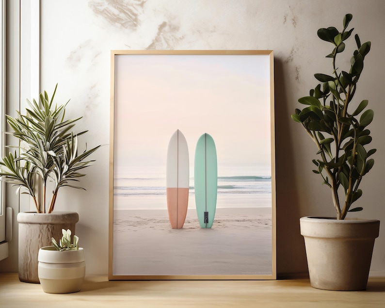 May include: A framed print of two surfboards on a beach. The surfboards are white and light blue with a pink and green stripe. The beach is sandy and the water is blue.