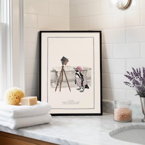 May include: Black framed art print featuring a penguin in a striped shirt and pink hat, standing near a bathtub with a vintage camera on a tripod. The print includes text at the bottom. The art is displayed in a bathroom setting.