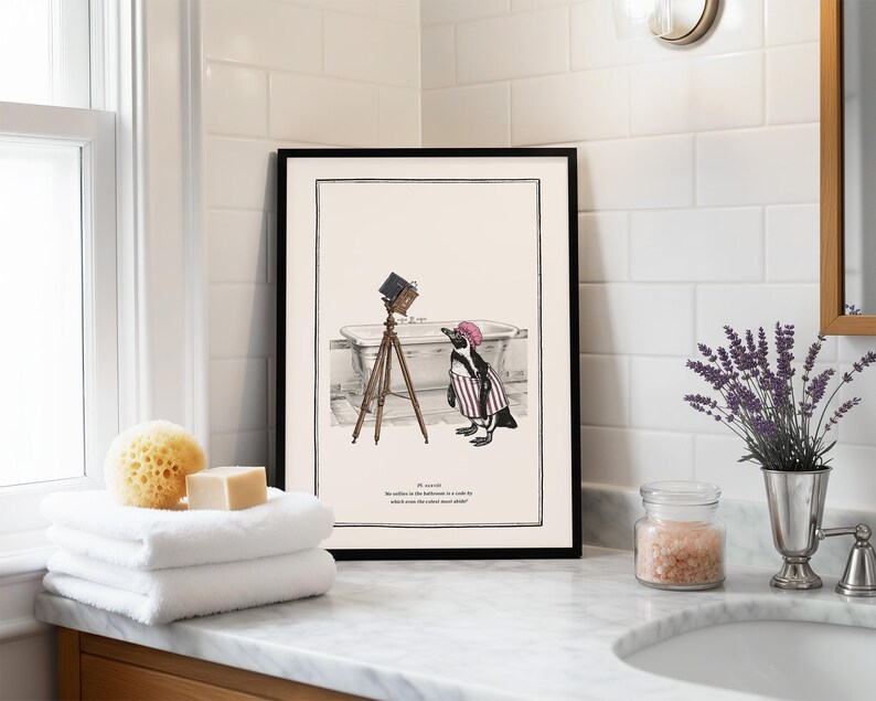 May include: Black framed art print featuring a penguin in a striped shirt and pink hat, standing near a bathtub with a vintage camera on a tripod. The print includes text at the bottom. The art is displayed in a bathroom setting.