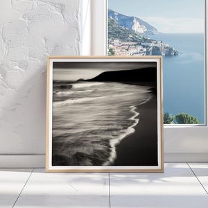 May include: A framed black and white photograph of a beach with waves crashing on the shore. The photo is in a wooden frame and is hanging on a white wall.