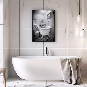 May include: A black and white photograph of a person in a bathtub. The person is wearing a hat and is sitting in a white bathtub with a shower head above it. The photo is framed and hanging on a white tiled wall.