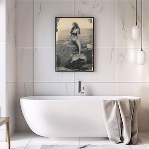 May include: A black and white photograph of a mermaid sitting on a rock in the ocean. The mermaid is wearing a seashell bra and has long flowing hair. Her tail is visible behind her.
