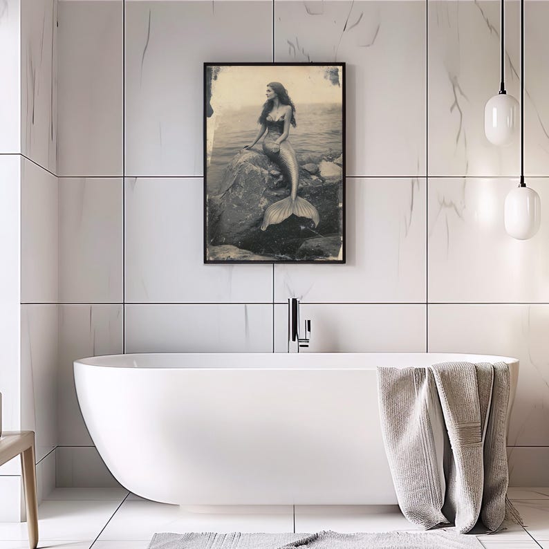 May include: A black and white photograph of a mermaid sitting on a rock in the ocean. The mermaid is wearing a seashell bra and has long flowing hair. Her tail is visible behind her.