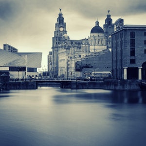 May include: A sepia-toned cityscape featuring historic buildings along a waterfront. Prominent structures include a clock tower, domed buildings, and a modern glass structure. The water in the foreground is smooth, reflecting the sky and buildings. A bridge spans the water.