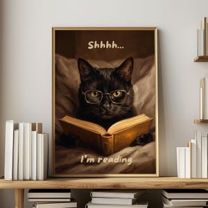 Puede incluir: Impresión enmarcada de un gato negro con gafas leyendo un libro. El texto "Shhhh..." y "I'm reading" están incluidos. La obra de arte tiene una paleta de colores cálidos, con el gato acostado en una cama.