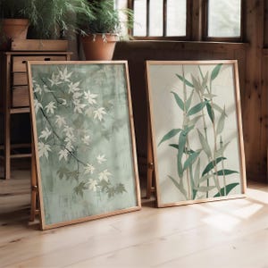 May include: Two framed art prints with wooden frames. One print features a branch with white maple leaves against a muted green background. The other print displays bamboo stalks and leaves in shades of green and brown against a light background.