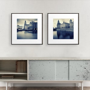 May include: Two framed prints of a cityscape in sepia tones. The images depict buildings along a body of water, with a cloudy sky overhead. The frames are black, and the prints are displayed on a light-colored cabinet with sliding doors.