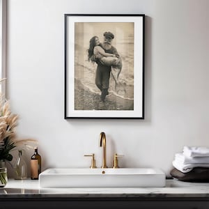 K&ouml;nnte beinhalten: Ein Schwarzwei&szlig;-Foto aus der Zeit, das einen Mann in einer Matrosenm&uuml;tze zeigt, der eine Meerjungfrau in seinen Armen h&auml;lt. Das Foto ist in einem schwarzen Rahmen gerahmt und h&auml;ngt an einer wei&szlig;en Wand.