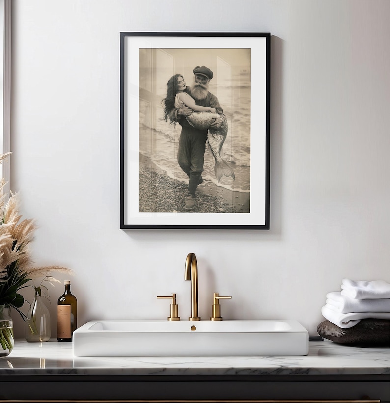 K&ouml;nnte beinhalten: Ein Schwarzwei&szlig;-Foto aus der Zeit, das einen Mann in einer Matrosenm&uuml;tze zeigt, der eine Meerjungfrau in seinen Armen h&auml;lt. Das Foto ist in einem schwarzen Rahmen gerahmt und h&auml;ngt an einer wei&szlig;en Wand.
