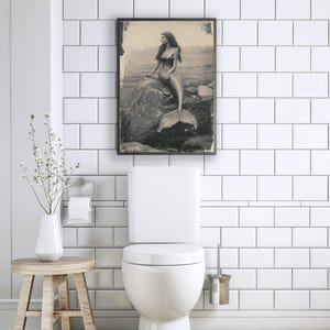 May include: A black and white photo of a mermaid sitting on a rock in the ocean. The photo is framed and hanging on a white tiled wall in a bathroom.