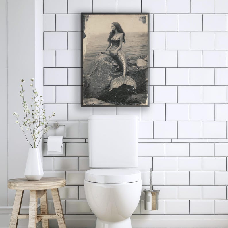 May include: A black and white photo of a mermaid sitting on a rock in the ocean. The photo is framed and hanging on a white tiled wall in a bathroom.