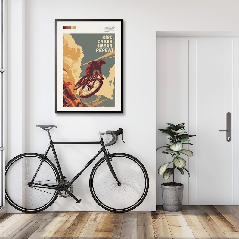 May include: A framed poster with the text "RIDE, CRASH, SWEAR, REPEAT" and an illustration of a mountain biker. A black road bike is leaning against the wall. A potted plant sits next to a white door.