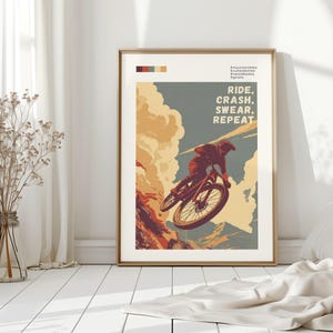 May include: Framed art print featuring a mountain biker mid-air. The artwork has a vintage style with a color palette of orange, blue, and beige. Text reads "RIDE, CRASH, SWEAR, REPEAT". The print is displayed in a room with white walls and a wooden floor.