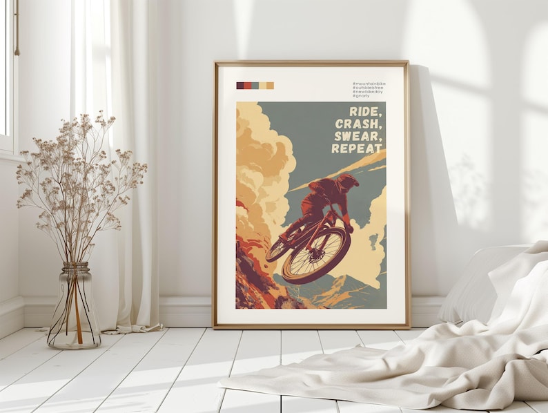 May include: Framed art print featuring a mountain biker mid-air. The artwork has a vintage style with a color palette of orange, blue, and beige. Text reads "RIDE, CRASH, SWEAR, REPEAT". The print is displayed in a room with white walls and a wooden floor.