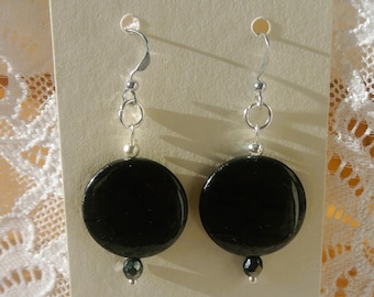 May include: A pair of black disc earrings with silver-colored hooks and accents. Each earring features a large, round, glossy black bead, a small silver bead, and a dark gray bead at the bottom. The earrings are displayed on a cream-colored card.
