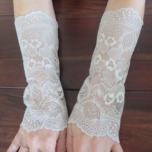May include: Two beige lace arm cuffs with floral patterns. The cuffs are made of delicate lace and are designed to be worn on the forearms. The cuffs are a neutral color and have a scalloped edge.