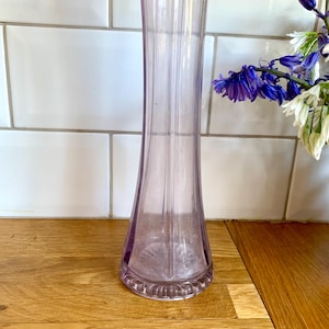 May include: A tall, slender, lilac-coloured glass vase with vertical ridges. The vase has a flared base and a decorative rim. The background includes white tiles and a wooden surface. Blue and white flowers are visible.