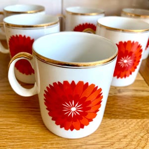 May include: Set of white ceramic mugs with a vibrant red and orange floral design. Each mug features a gold rim and handle, adding a touch of elegance. The mugs are arranged on a wooden surface, showcasing their decorative appeal.