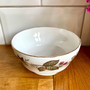 Puede incluir: Un cuenco de cerámica blanca con borde dorado y diseño floral con hojas verdes y bayas rojas. El cuenco está sobre una superficie de madera, con un fondo de azulejos blancos.