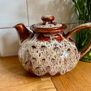 Puede incluir: Una tetera de cerámica marrón y blanca con tapa y asa. La tetera tiene un patrón moteado y un acabado brillante. El pico y el asa son de un tono marrón más oscuro. Un artículo decorativo para la cocina o el comedor.