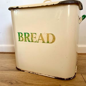 Op de afbeelding: Crèmekleurige broodtrommel met het woord "BREAD" in groene en gouden letters. De trommel heeft een gebogen handvat op het deksel en groene handvatten aan de zijkanten. De randen vertonen slijtage, wat het een vintage uitstraling geeft.