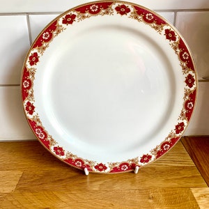 May include: A white ceramic dinner plate with a red and gold floral border. The plate is on a wooden surface.