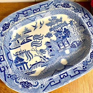 May include: An oval ceramic serving platter featuring a blue and white willow pattern. The design includes pagodas, a bridge, willow trees, and two birds in flight. The wide border displays a geometric fretwork pattern with floral accents.