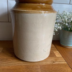 Puede incluir: Un tarro de gres vintage con cuerpo beige y borde vidriado marrón. El recipiente cilíndrico tiene una abertura ancha y una forma ligeramente cónica. El tarro se utiliza probablemente para el almacenamiento de alimentos.