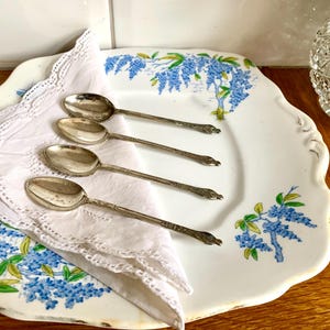 Può includere: Quattro cucchiai d'argento con manici decorati sono disposti su un tovagliolo di pizzo bianco, posto su un piatto decorativo. Il piatto presenta un motivo floreale blu. La scena è ambientata su una superficie di legno.