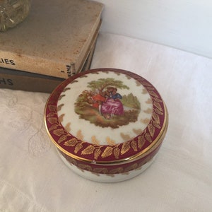 May include: A round porcelain trinket box with a red and gold border. The lid features a painted scene of two people in a garden setting.