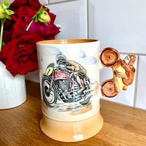 Puede incluir: Una taza de cerámica con un diseño de motocicleta marrón y blanco. La taza tiene un asa marrón con forma de rueda de motocicleta.