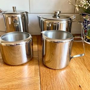 Può includere: Un set di quattro articoli da cucina in acciaio inossidabile che include due teiere con coperchi e manici, una zuccheriera e una lattiera con manico. Ogni pezzo presenta una finitura argentata lucida e riflettente e un design cilindrico liscio.