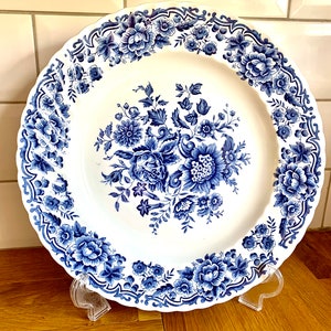 Peut inclure: Une assiette en céramique blanche avec un motif floral bleu. L'assiette a un bord festonné et est décorée d'un grand motif floral au centre.