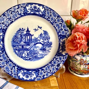 Puede incluir: Un plato de porcelana azul y blanco con un borde floral y un diseño central de una pagoda y un puente en un jardín.