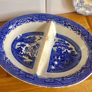 May include: Oval divided serving dish with a blue and white Willow pattern. The dish is divided into two sections by a raised divider. The rim and the outer edges of the dish have a detailed blue border. The dish is on a wooden surface.
