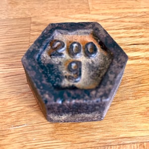 May include: A hexagonal cast iron weight with the number 200 and the letter g on it. The weight is rusty and has a dark brown colour.