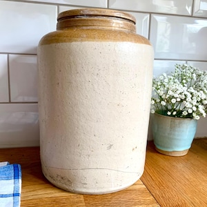 May include: A large, antique stoneware crock with a lid. The crock is cylindrical with a light beige body and a darker tan rim and lid. A crack runs horizontally across the lower portion of the crock. A small blue and white vase with white flowers is to the right.