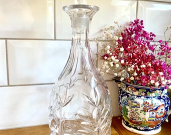 May include: A clear, cut-glass decanter with a faceted stopper. The decanter features a detailed leaf and starburst pattern. The stopper is a geometric shape. The decanter is on a wooden surface.