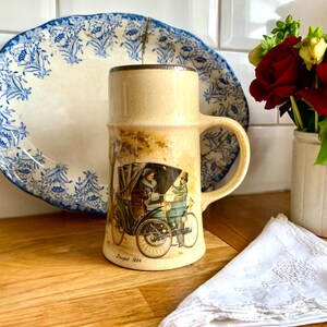 May include: A ceramic mug with a brown rim and handle. The mug features a vintage illustration of a car with two people inside. The text "Peugeot 1904" is printed on the bottom of the mug.