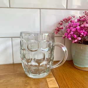 Op de afbeelding: Een heldere glazen bierpul met een handvat en een gestructureerd oppervlak. De pul staat op een houten oppervlak, met een wit betegelde muur op de achtergrond. Rechts van de pul staat een kleine pot met roze bloemen.